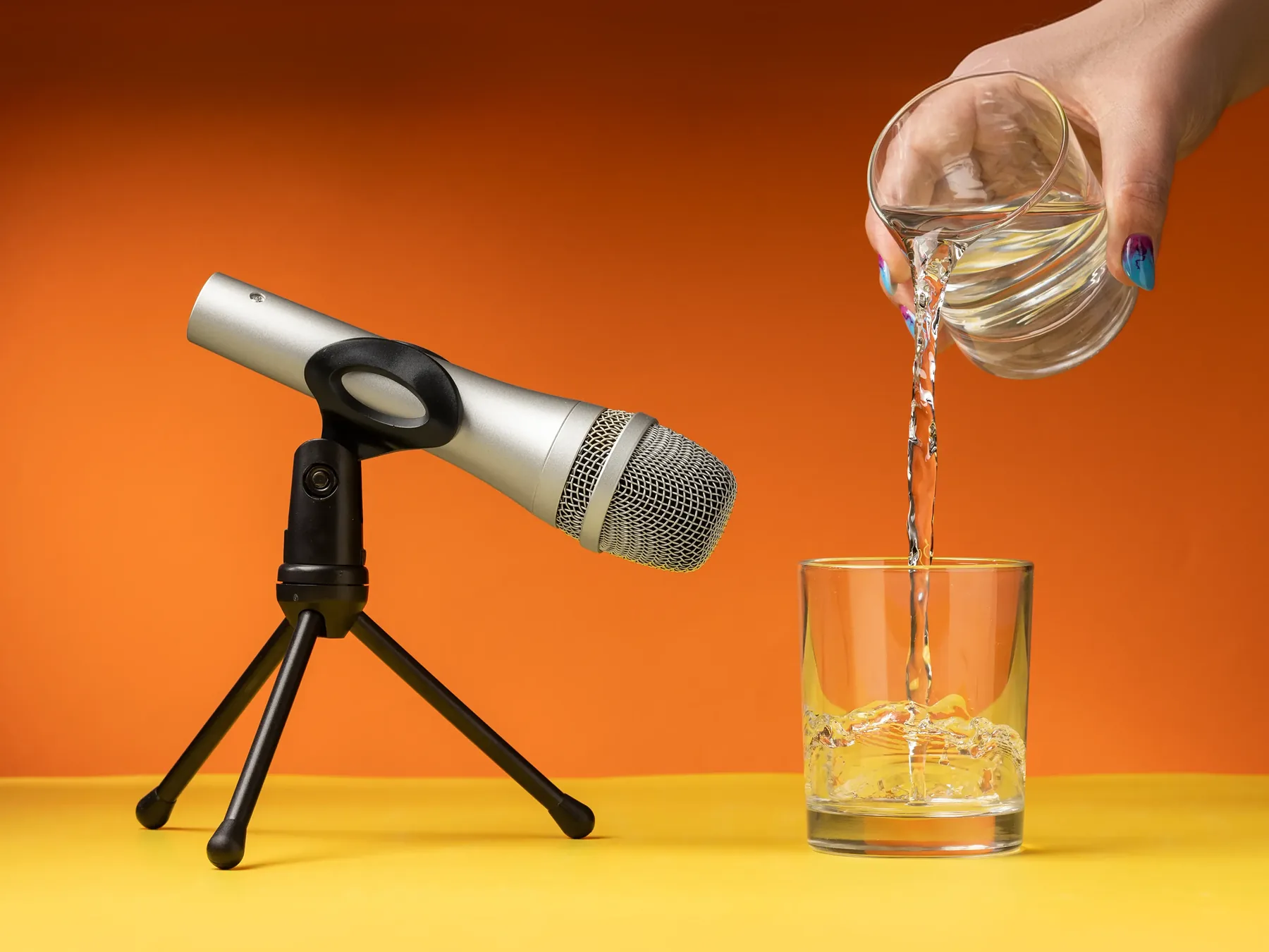 A microphone on a desk tripod beside a glass of water being poured, against an orange and yellow backdrop