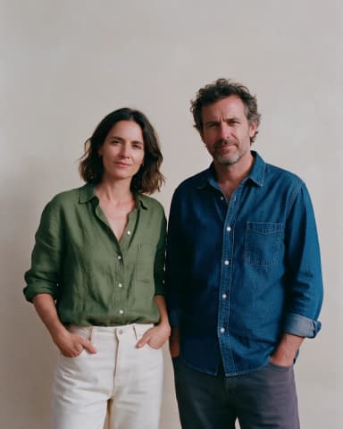 Mette Holm and Jonas Kjær standing side by side against a plain sand backdrop