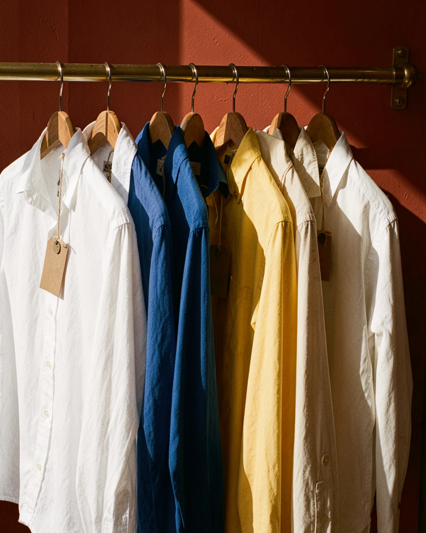 A rail of finished shirts in white, cobalt, yellow, and ecru against a terracotta wall