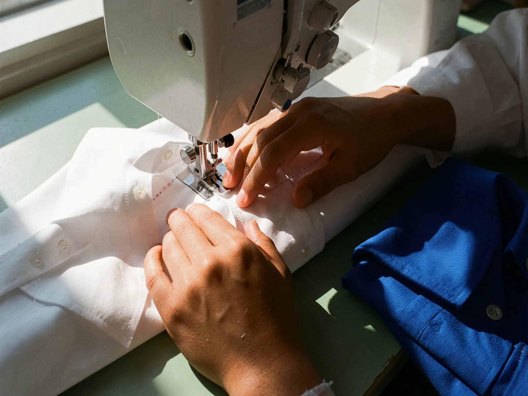 Hands guiding white poplin under a sewing machine foot along a shirt collar