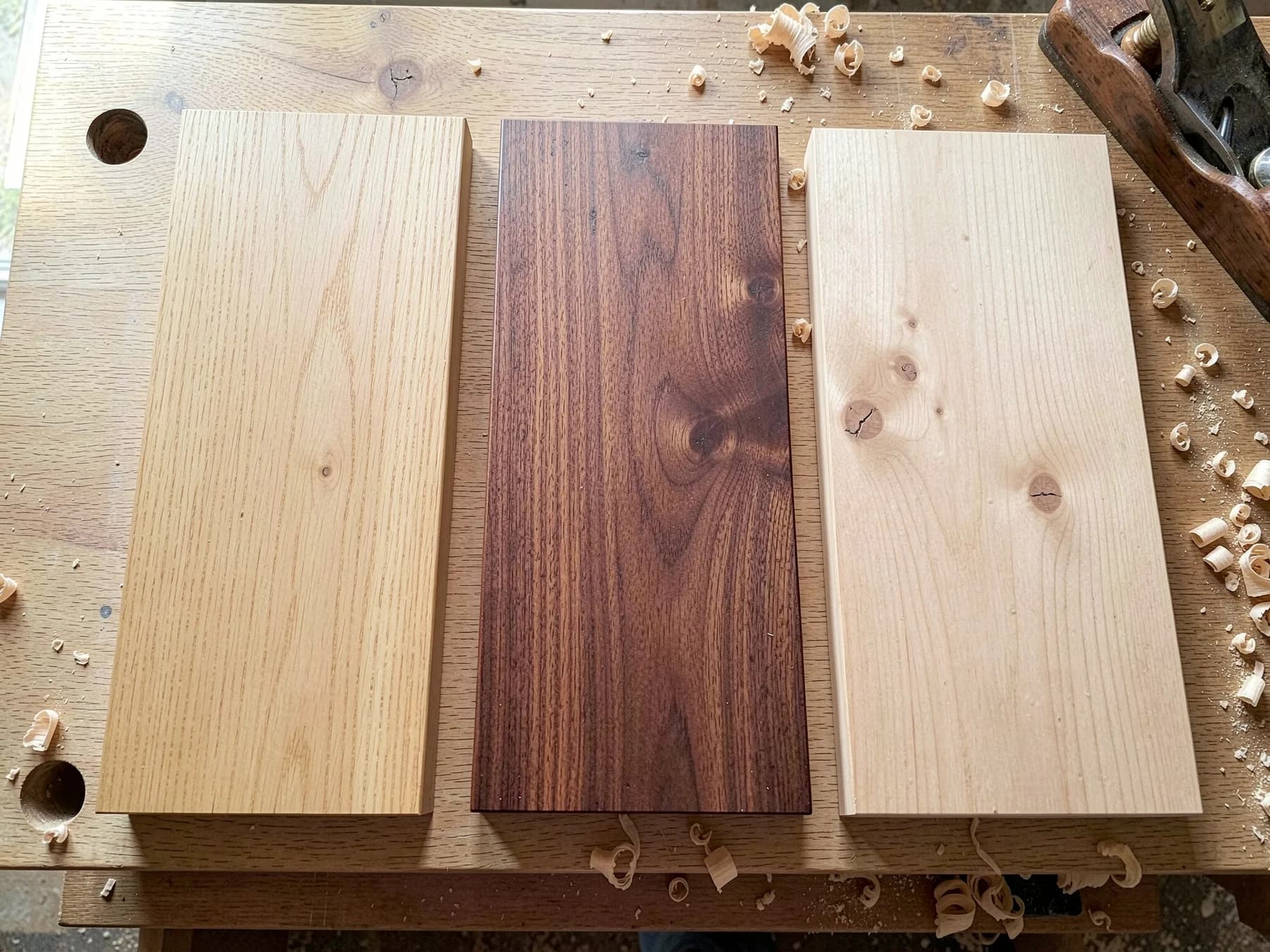 3 oiled boards side by side on a workshop bench: pale oak, dark walnut, and light ash, with shavings and a hand plane